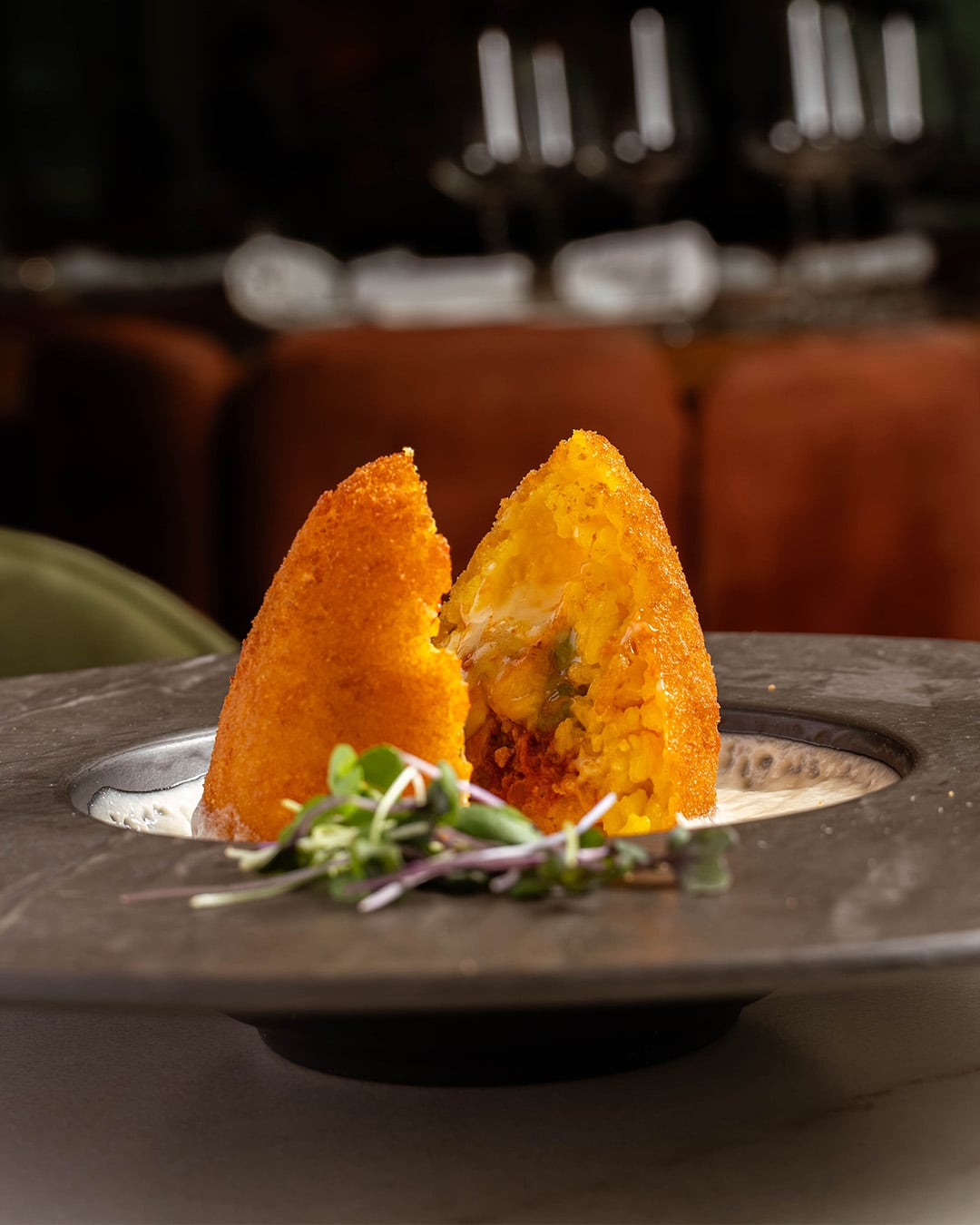 Arancino siciliano partido por la mitad sobre un plato oscuro, con microhojas