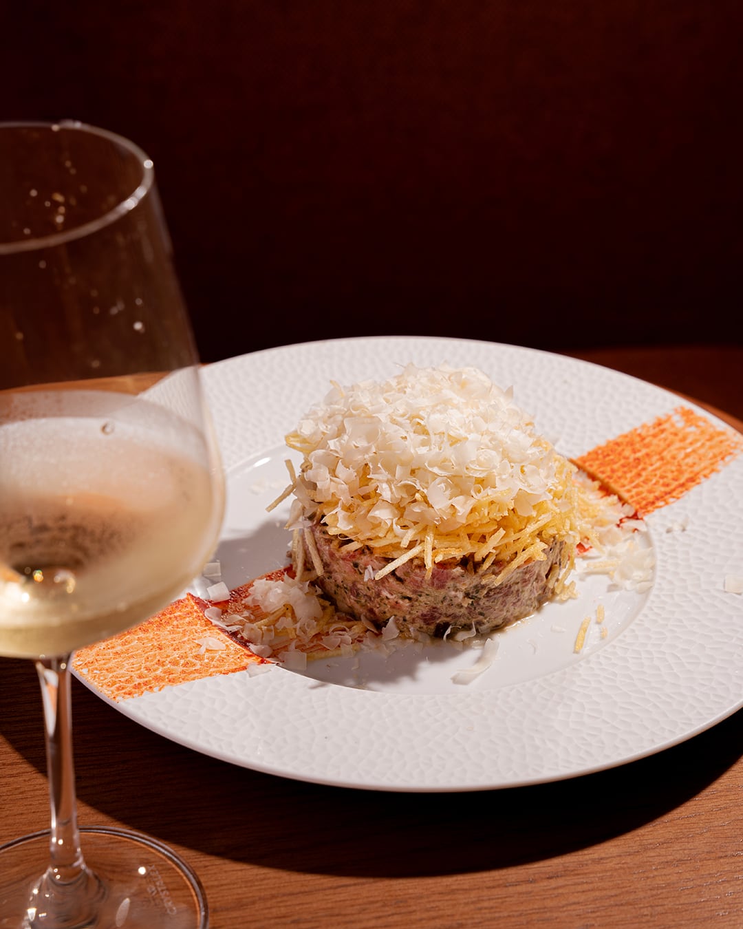 Steak tartare con queso rallado sobre un plato blanco, junto a una copa de vino blanco